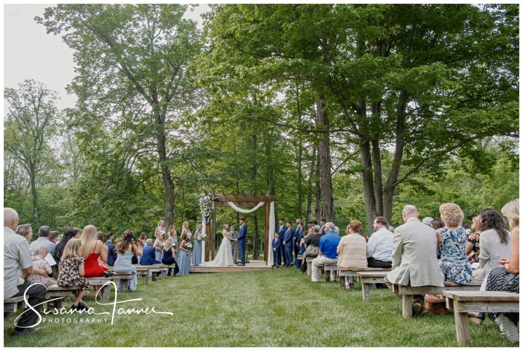 Laurel Mill Barn Wedding, Bloomington IN, wedding ceremony outside under the trees with guests sitting on wooden benches