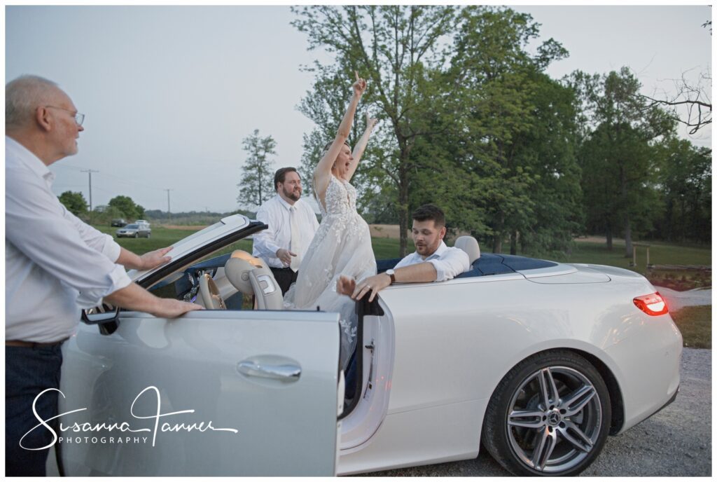 Bride standing with her arms in in white mercedes car as they drive away at end of night